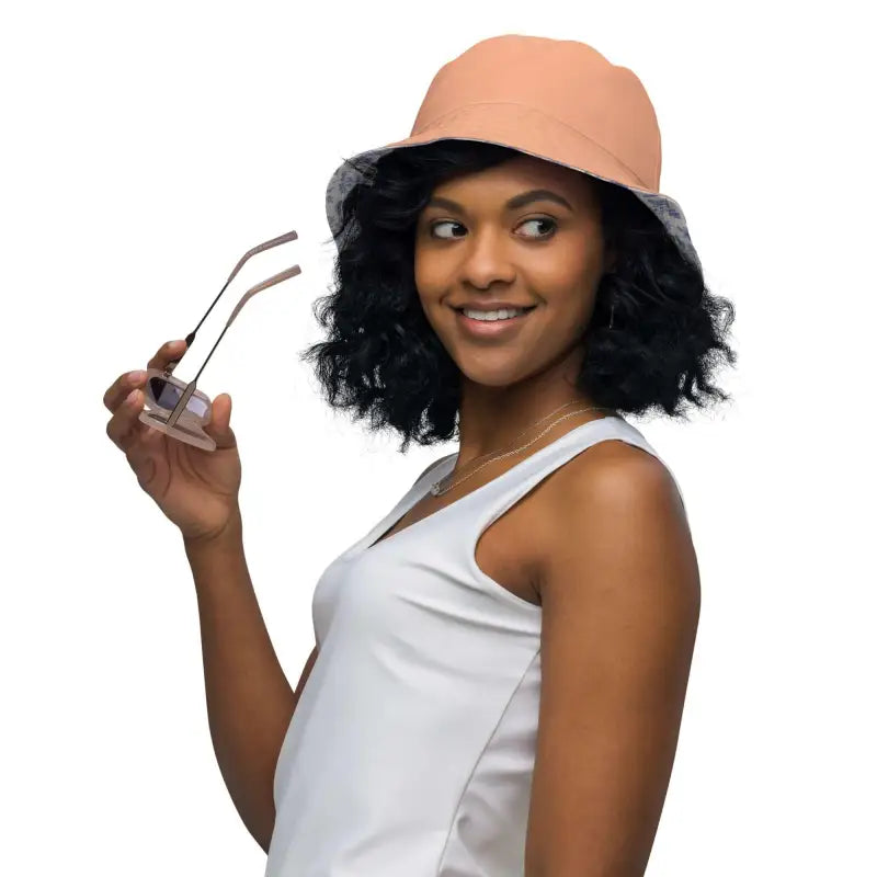 Smiling woman in a solid peach reversible bucket hat and white tank top holding sunglasses
